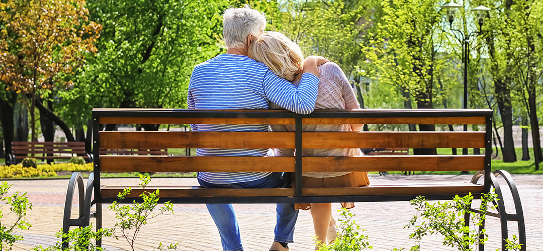 A mature couple sits on a park bench on a sunny day